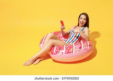 Full body length happy young woman wear red blue swimsuit sit on inflatable ring hold mobile cell phone isolated on vivid yellow color wall background studio Summer hotel pool sea rest sun tan concept
