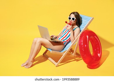 Full Body Length Happy Young Woman Wear Red Blue One-piece Swimsuit Sit On Chair Work Hold Laptop Pc Isolated Over Vivid Yellow Color Wall Background Studio. Summer Hotel Pool Sea Rest Sun Tan Concept