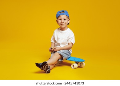 Full body length joyful kid boy wearing trendy blue cap and white t-shirt, smiling while sitting on modern skateboard against yellow background. - Powered by Shutterstock