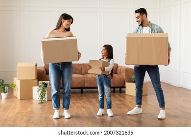 Full Body Length Portrait Of Cheerful Family Of Three People Housing, Happy Smiling Man, Woman And Little Girl Holding And Carrying Cardboard Boxes Standing In Empty Living Room. House Move Concept