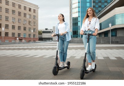 Full body length shot of two best female friends driving black motorized kick scooters in the city centre, smiling casual young ladies spending free time together on weekend, enjoying ride