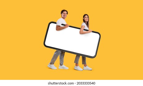 Full body length side view profile of cheerful couple carrying big smart phone with blank white screen, walking with gadget under arm, looking at camera isolated on yellow studio background, mock up - Shutterstock ID 2033333540