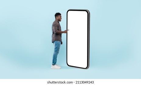 Full Body Length Side View Of Black Man Using Big Smartphone With Blank White Screen Touching Huge Display Panel With Finger, Cheerful Guy Standing On Blue Background, Ordering Food Delivery, Mock Up