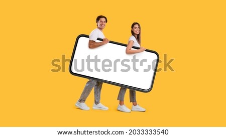 Full body length side view profile of cheerful couple carrying big smart phone with blank white screen, walking with gadget under arm, looking at camera isolated on yellow studio background, mock up