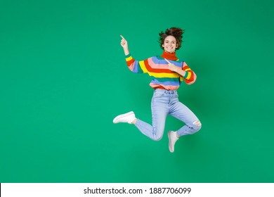 Full body length smiling laughing pretty young brunette woman 20s in colorful sweater jumping point index fingers aside on mock up copy space isolated on bright green color background studio portrait