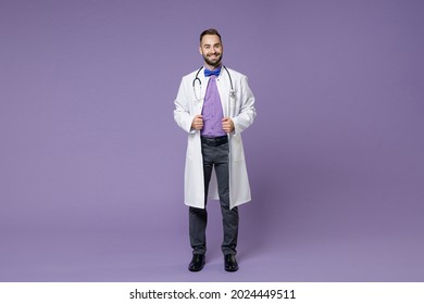 Full Body Length Smiling Young Bearded Doctor Man In White Medical Gown Standing Looking Camera Isolated On Violet Colour Wall Background Studio Portrait. Healthcare Personnel Health Medicine Concept