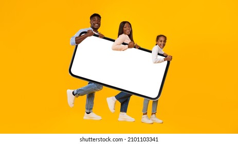 Full Body Length Of Three Cheerful Black People Carrying Big Cell Phone With Blank White Screen, Walking With Gadget Under Arm, Looking At Camera Isolated On Yellow Orange Studio Background, Mock Up