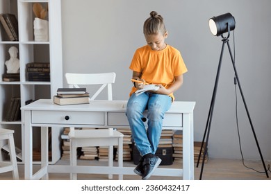 Full body lifestyle cheeky boy schoolboy 7-8 children in casual clothes sits on a table in the modern school library and does homework, writing in notebook - Powered by Shutterstock