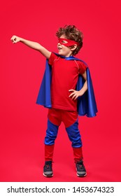 Full Body Little Boy In Mask And Cape Holding Fist Forward Standing In Superhero Pose On Red Background