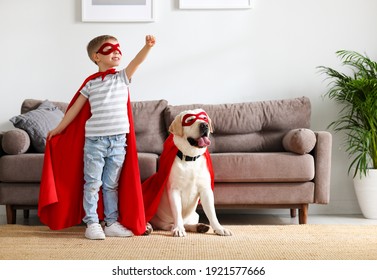 Full body of little boy in red superhero cloak and mask raising hand while playing with funny dog dressed in similar costume in living room at home