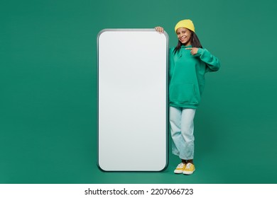 Full body little kid teen girl of African American ethnicity 13-14 years old wear casual hoody hat point finger on big blank screen mobile cell phone with area isolated on plain dark green background - Shutterstock ID 2207066723