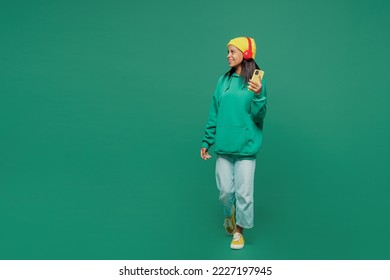 Full body little kid teen girl of African American ethnicity 13-14 year old in casual hoody hat headphones listen music use mobile cell phone isolated on plain dark green background Childhood concept - Powered by Shutterstock