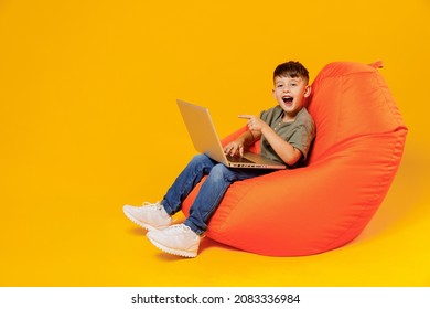 Full Body Little Small Fun Boy 6-7 Years Old In Green T-shirt Sit In Bag Chair Hold Use Work Point On Laptop Pc Computer Isolated On Plain Yellow Background Mother's Day Love Family Lifestyle Concept