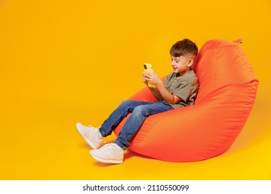 Full body little small happy boy 6-7 years old wearing green t-shirt sit in bag chair hold in hand use mobile cell phone isolated on plain yellow background. Mother's Day love family lifestyle concept