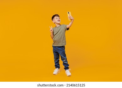 Full body little small happy boy 6-7 years old in green t-shirt do selfie shot on mobile cell phone show v-sign gesture isolated on plain yellow background. Mother's Day love family lifestyle concept