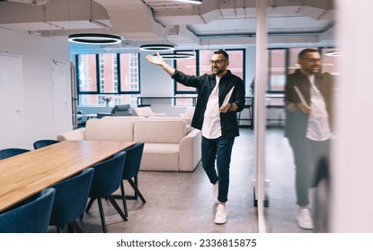 Full body of male entrepreneur in smart casual outfit spreading arms welcoming while walking in modern workspace near glass wall with laptop - Shutterstock ID 2336815875
