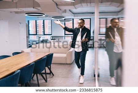 Full body of male entrepreneur in smart casual outfit spreading arms welcoming while walking in modern workspace near glass wall with laptop