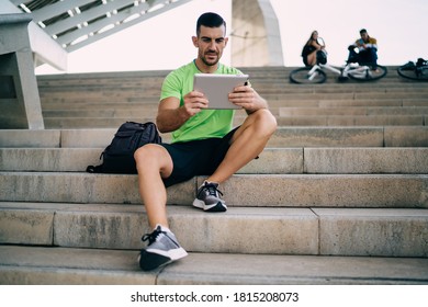 Full body of mature ethnic male in summer sportswear and sneakers with backpack using tablet while resting after workout on stairs under contemporary construction - Powered by Shutterstock