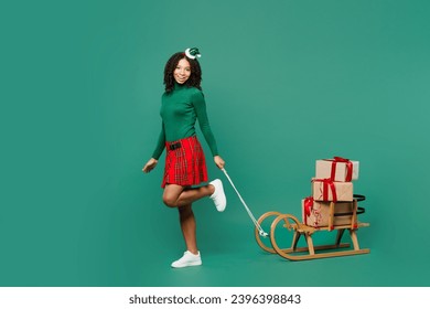 Full body merry little kid teen girl wear hat casual clothes posing carry on sled stack of gift present boxes isolated on plain green background. Happy New Year celebration Christmas holiday concept - Shutterstock ID 2396398843