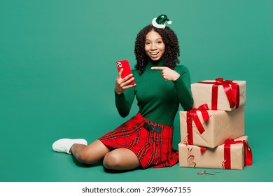 Full body merry little kid teen girl wear hat casual clothes sit hold mobile cell phone present boxes with gift ribbon bow posing isolated on plain green background. Happy New Year Christmas concept - Powered by Shutterstock