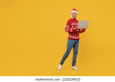 Full body merry young IT man wear red knitted Christmas sweater Santa hat posing look aside hold use work on laptop pc computer isolated on plain yellow background. Happy New Year 2023 holiday concept - Powered by Shutterstock