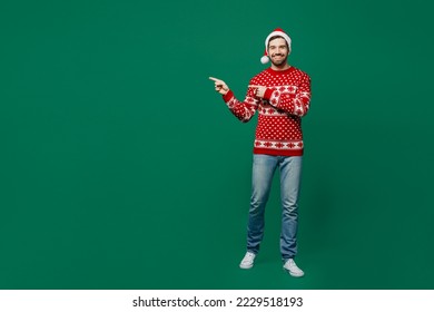 Full body merry young man 20s wear red warm knitted sweater Santa hat posing point index finger aside on workspace area isolated on plain dark green background studio. New Year 2023 holiday concept - Powered by Shutterstock