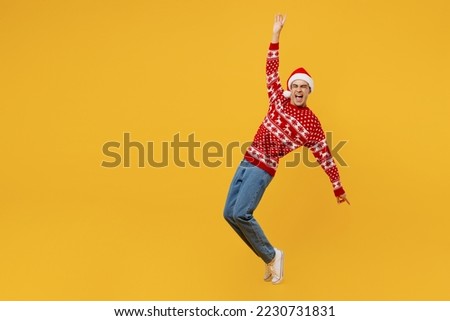 Full body merry young man wear red knitted Christmas sweater Santa hat posing leaning back with outstretched hands stand on toes isolated on plain yellow background Happy New Year 2023 holiday concept