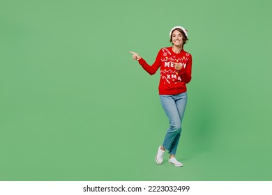 Full body merry young woman wear knitted xmas sweater Santa hat posing point indicate on workspace area isolated on plain pastel light green background. Happy New Year 2023 celebration holiday concept