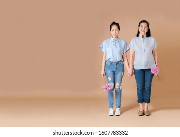 Full Body Of An Middle Age Asian Woman And A Young Asian Woman, They Holding Hands On Beige Background. They Are Smiling And Holding Pink Flower In Hands. With Copy Space Area.