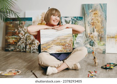 Full body minded elderly artist woman 50 years old wearing casual clothes sit on floor hold in hand show painting on canvas artwork spend free spare time in living room indoor. Leisure hobby concept - Powered by Shutterstock