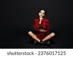 Full body minded young woman with Halloween makeup face art mask wearing clown costume red dress sit hold use mobile cell phone isolated on plain black background studio. Scary holiday party concept