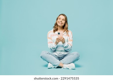 Full body minded young woman wears striped hoody sitting hold in hand use mobile cell phone look aside on area isolated on plain pastel light blue cyan background studio portrait. Lifestyle concept - Powered by Shutterstock