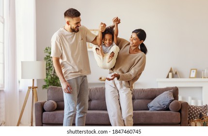 Full body multiethnic father and mother smiling and lifting mixed race son near couch in cozy living room