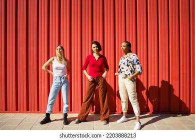 Full Body Of Multiethnic Stylish Female Friends Standing With Hands In Pockets Near Red Wall On Sunny Street Of City