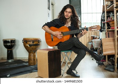 Full Body Music Shot Of A Flamenco Styled Young Long Haired Musician Passionately Playing The Spanish Acoustic Guitar On A Humble Rehearsal Studio With More Percussion Instruments.