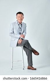 Full Body Of An Old Asian Man Wear Light Grey Suit Sitting On A Grey Chair And Cross One's Legs In Studio White Background. Isolated Picture Of Happiness Asian Old Man.