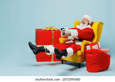 Full body old Santa Claus man in Christmas hat red suit sit in armchair near gift bag play pc game joystick console isolated on plain blue background studio. Happy New Year 2022 merry ho x-mas concept