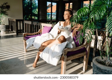 Full body of optimistic female traveler smiling and relaxing on bench with cup of hot drink on tropical resort in sunrise - Shutterstock ID 2300580379