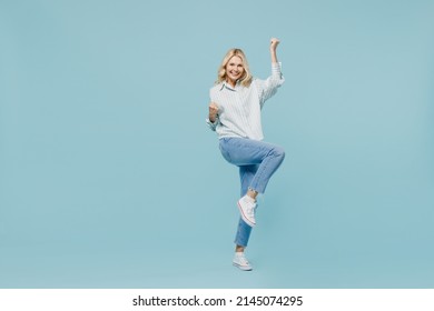 Full body overjoyed elderly woman 50s in casual striped shirt do winner gesture clench fist raise up leg isolated on plain pastel light blue color background studio portrait. People lifestyle concept