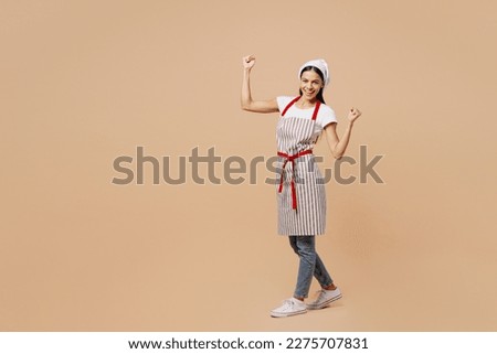 Full body overjoyed satisfied sideways young housewife housekeeper chef baker latin woman wear apron toque hat walk do winner gesture isolated on plain pastel light beige background. Cook food concept