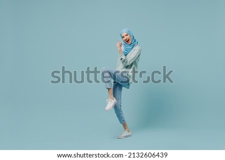 Full body overjoyed young arabian asian muslim woman in abaya hijab do winner gesture clench fist isolated on plain blue background studio portrait. People uae middle eastern islam religious concept