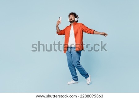 Full body overjoyed young fun Indian man wears orange red shirt white t-shirt headphones listen to music use mobile phone isolated on plain pastel light blue cyan background studio. Lifestyle concept