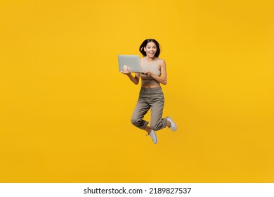 Full Body Overjoyed Young Latin Woman 30s She Wear Basic Beige Tank Shirt Hold Use Work On Laptop Pc Computer Jump High Lok Camera Isolated On Plain Yellow Backround Studio. People Lifestyle Concept