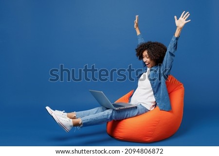 Full body overjoyed young smiling student happy black woman in casual clothes shirt white tshirt sit in bag chair hold use work on laptop pc computer finish job isolated on plain dark blue background.