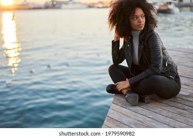 Full Body Of Pensive Young African American Female Tourist With Bouncy Hair In Stylish Black Coat And Jeans Thoughtfully Looking Away While Lounging On Pier Near Lake On Sunset