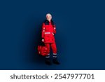 Full body photo of attractive senior woman ambulance worker hold first aid kit dressed red uniform isolated on dark blue color background