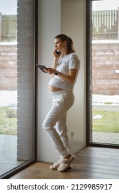 Full Body Photo Of Attractive Woman Making Phone Call With Tablet In Hand Being At Home During Pregnancy