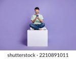 Full body photo of attractive young guy hold gadget sit white cube apple samsung wear trendy gray look isolated on violet color background