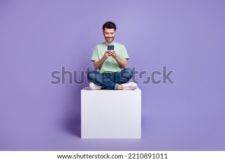 Full body photo of attractive young guy hold gadget sit white cube apple samsung wear trendy gray look isolated on violet color background