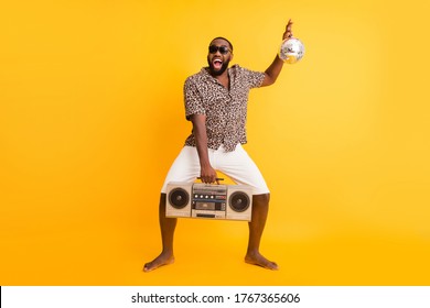 Full body photo of crazy funny dark skin guy hold retro tape recorder glitter disco ball rejoicing resting party wear shorts leopard shirt sun glasses isolated yellow background